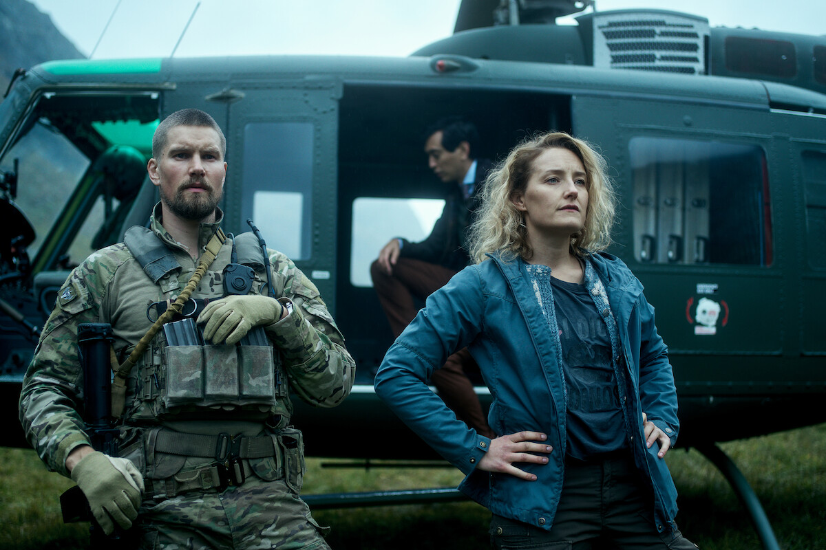 A soldier in camouflage and tactical gear stands confidently next to a determined woman in casual outdoor attire, with a helicopter and a seated man in the background, set in a rugged outdoor location.