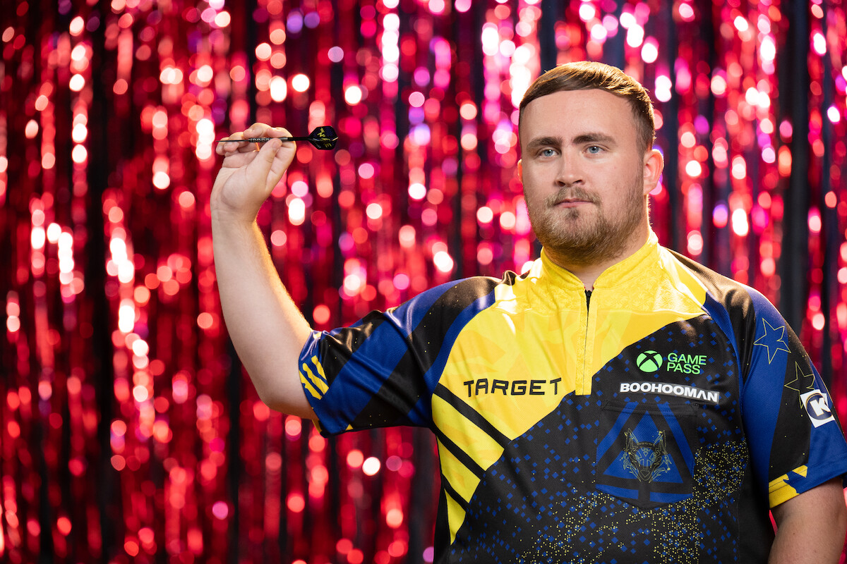 Man in a colorful dart shirt holding a dart, standing in front of a shiny red and pink tinsel curtain background, looking focused and ready to throw.