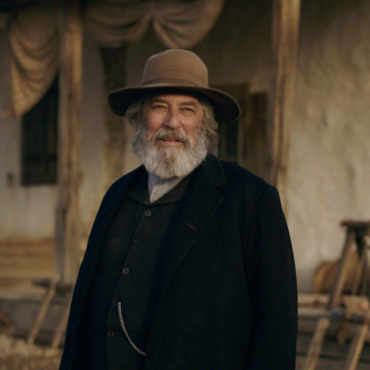 Ciarán Hinds as Samuel Hamilton with gray beard in old-fashioned dark clothes and wide-brimmed hat stands outside a rustic wooden building with draped curtains, suggesting a historical or western setting.