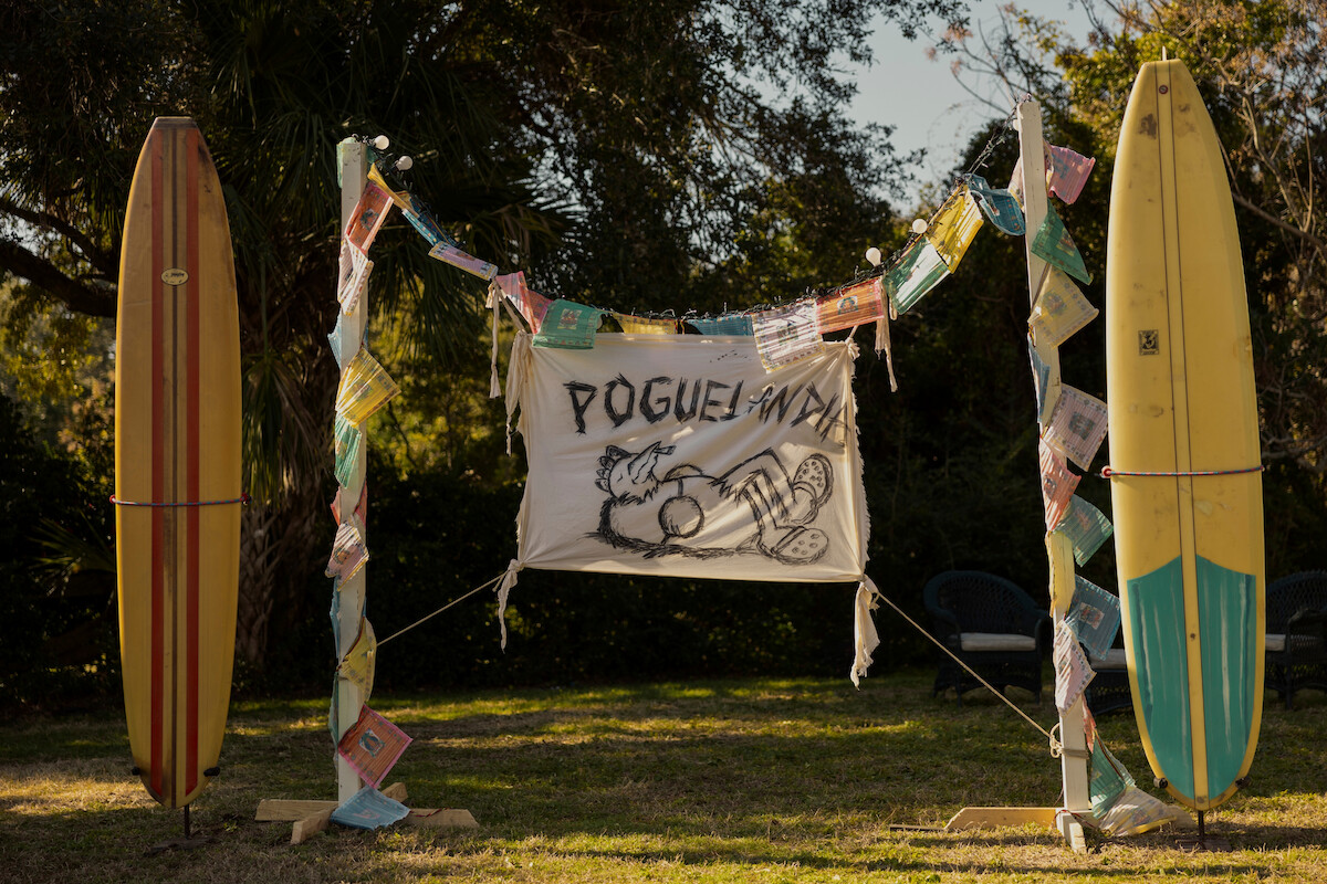 Two colorful surfboards stand upright outdoors with festive garlands and a hand-drawn “POGUELANDIA” banner between them, surrounded by trees and outdoor chairs on grass in a sunny backyard setting.
