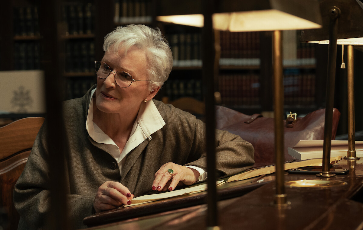 Elderly woman with short white hair and glasses sits at a wooden desk reading a book in a dimly lit library, surrounded by bookshelves, table lamps, and a leather bag.