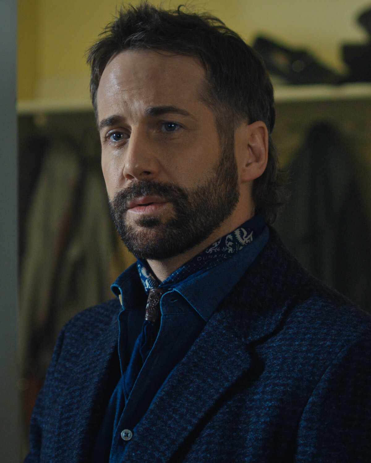 A man with a beard and mustache stands indoors in a dimly lit room, wearing a blue jacket and a scarf, with coats and shelves blurred in the background.