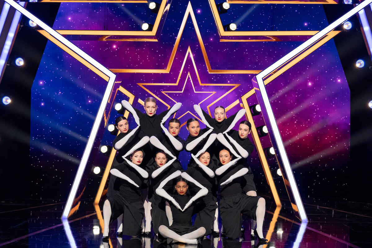 Ladymetry in black and white costumes performs a synchronized formation on a stage with a large illuminated star backdrop and vibrant purple and blue lighting effects.