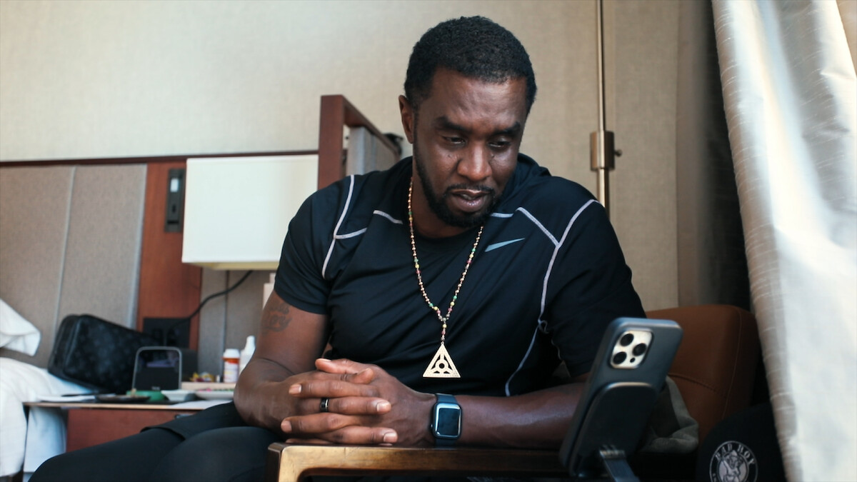 Man in black athletic wear sitting indoors, looking intently at a phone on a stand in a cozy, modern room with relaxed, thoughtful mood. Accessories include a smartwatch and pendant necklace.