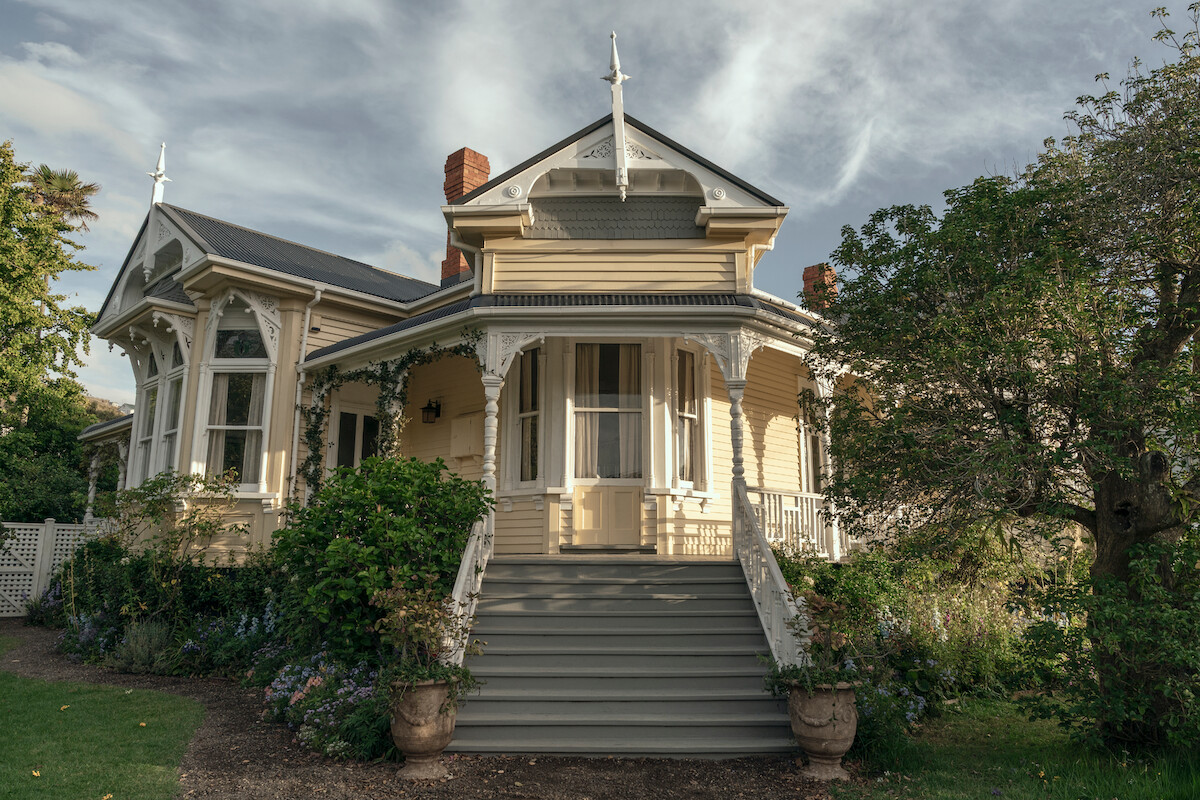 Victorian-style house with ornate trim, large porch, and front steps, surrounded by lush greenery and trees, under a partly cloudy sky in a calm, residential neighborhood.