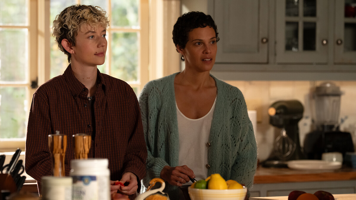Two people standing together in a cozy kitchen with a bowl of lemons, a mixer, and wooden utensils on the counter, sunlight coming through a window and cabinets in the background.