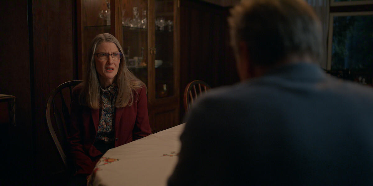 A woman with long gray hair and glasses sits at a dining table talking to a man in a dimly lit room with dark wood paneling and a glass cabinet in the background.