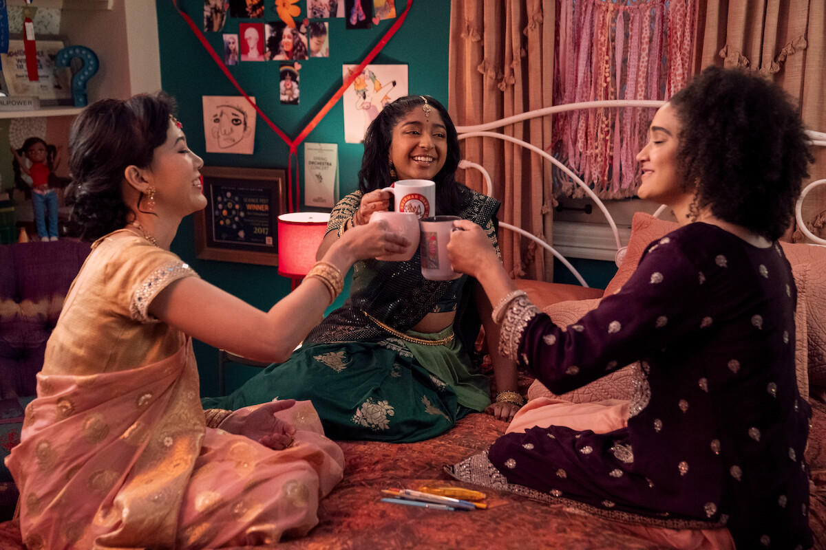 Ramona Young as Eleanor Wong, Maitreyi Ramakrishnan as Devi and Lee Rodriguez as Fabiola Torres sitting on a bed cheering mugs in ‘Never Have I Ever’ Season 4. 