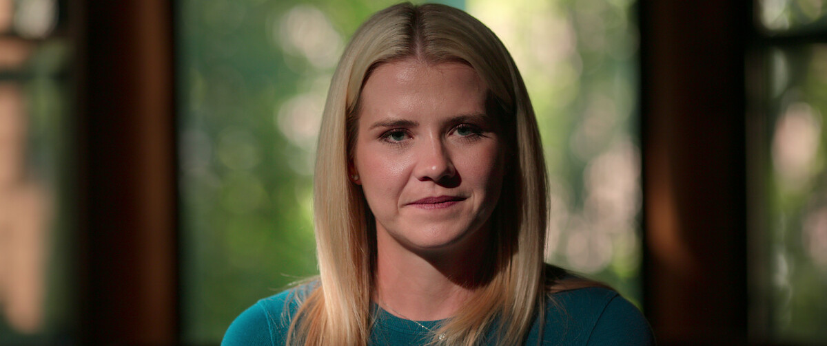 Blonde woman in a teal shirt sits indoors with soft natural light coming through windows behind her, creating a calm and introspective mood.