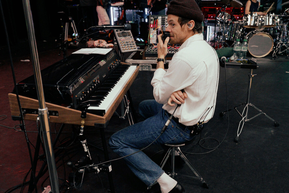 Harry Styles in casual clothes sits at a keyboard on stage, holding a microphone during a rehearsal. Musical equipment and other band members are visible in the background. The setting appears to be a live performance venue.