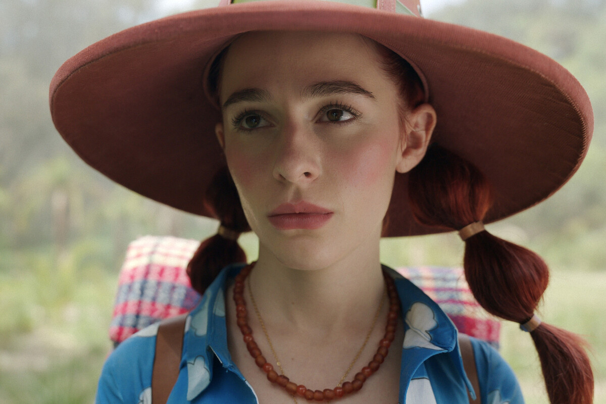Young woman with red hair in pigtails wearing a large brown hat, blue shirt with white spots, and a beaded necklace, standing outdoors with a backpack and a blurred green natural background.