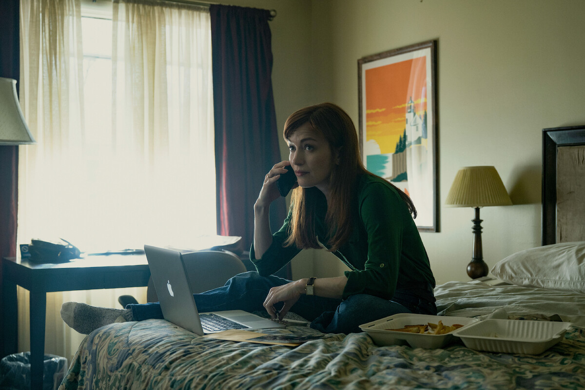 Woman sitting on a bed in a dimly lit room, talking on the phone, working on a laptop with papers and food containers nearby, window with light curtains and colorful wall art in the background.