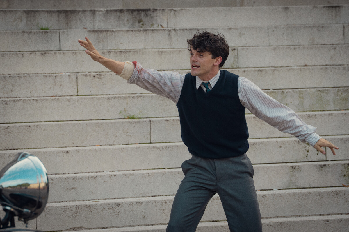 Young man in dress shirt, tie, and sweater vest stands on outdoor stone steps, arms outstretched and expression intense, with a motorcycle partially visible in the corner, suggesting a dramatic or urgent moment.