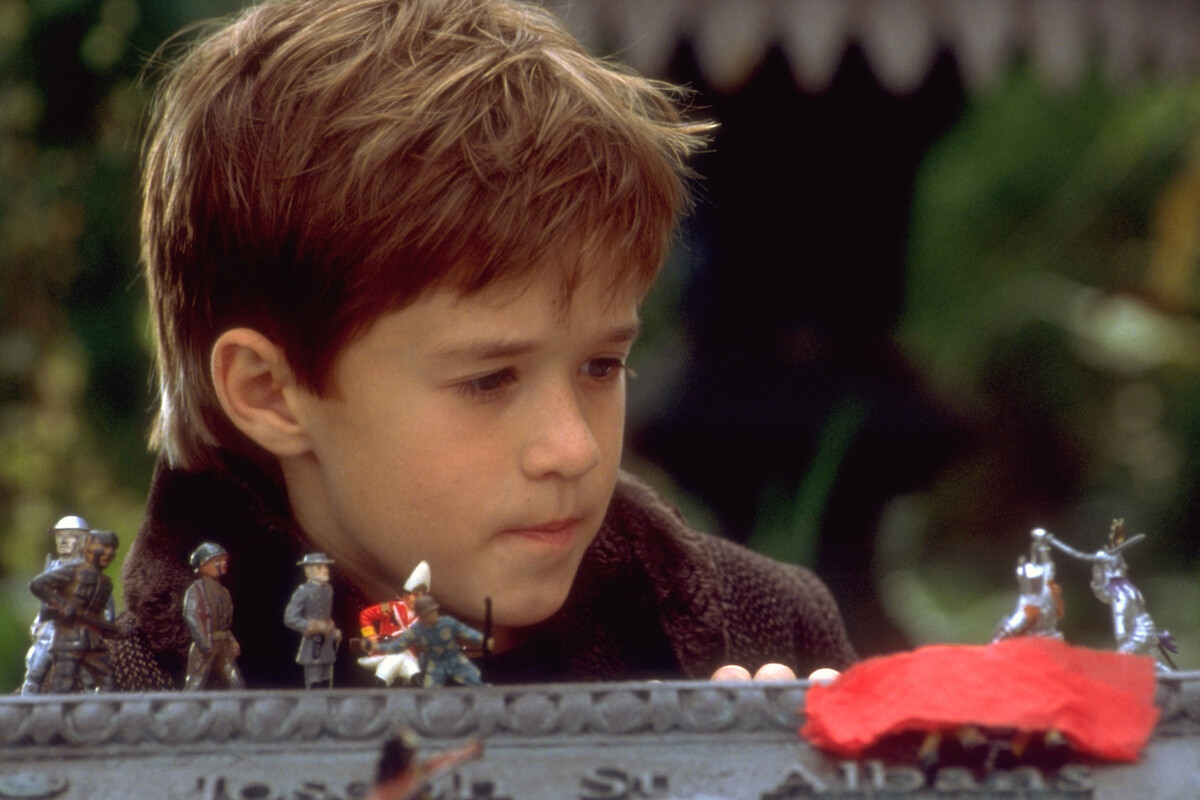 Young boy with brown hair looks thoughtfully at a collection of toy soldiers arranged in front of him outdoors, with greenery in the blurred background, suggesting a playful and imaginative setting.