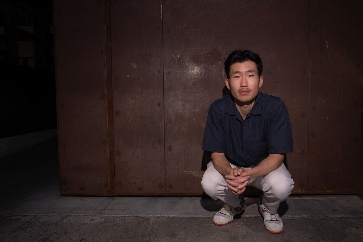 Man squatting on concrete in front of a large rusty metal wall at night, wearing a navy shirt, white pants, and sneakers, looking directly at the camera in an urban outdoor setting.