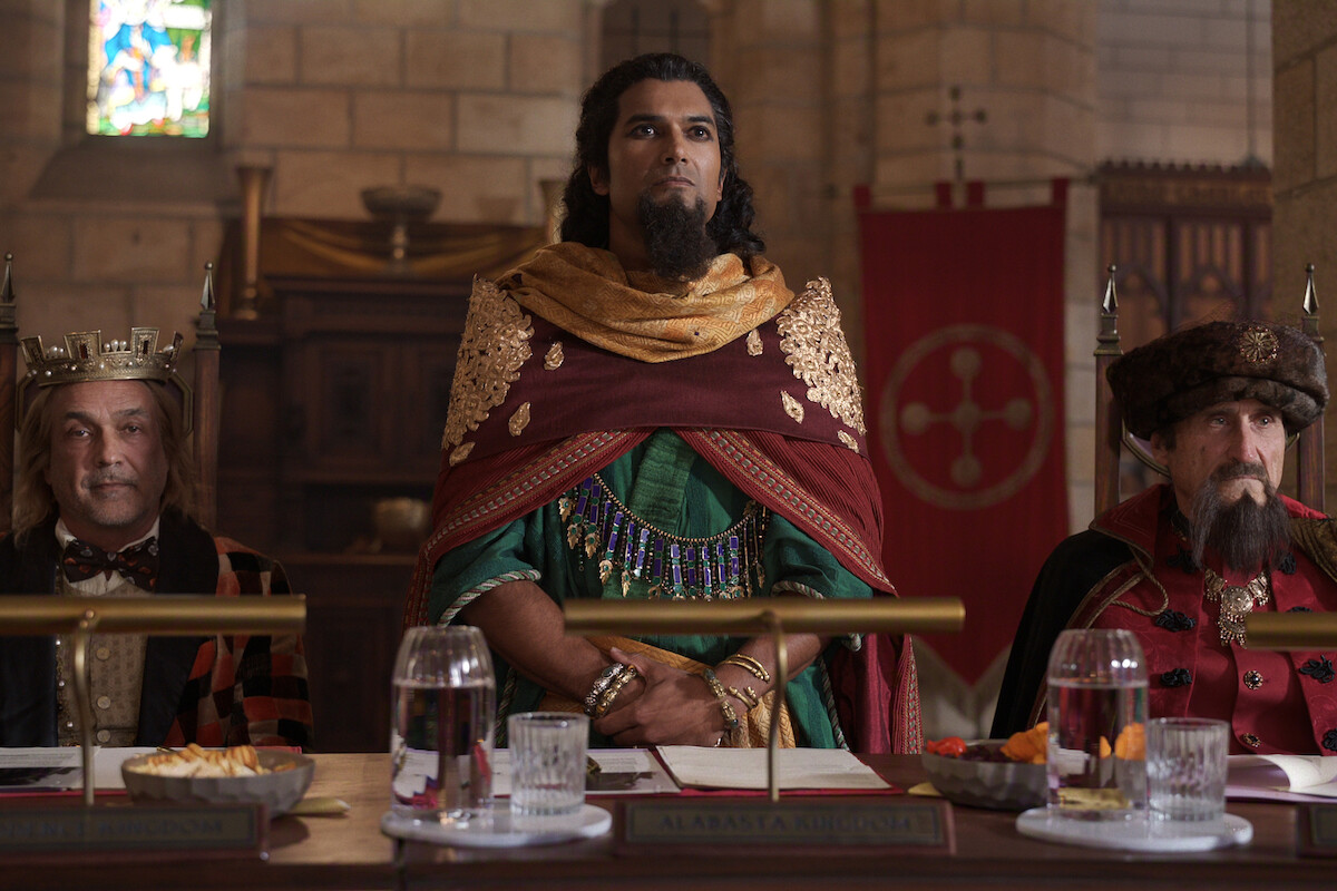 Three men in medieval royal attire sit at a table in a grand stone hall with stained glass windows, banners, and food and drinks set before them, evoking a historical or fantasy setting.