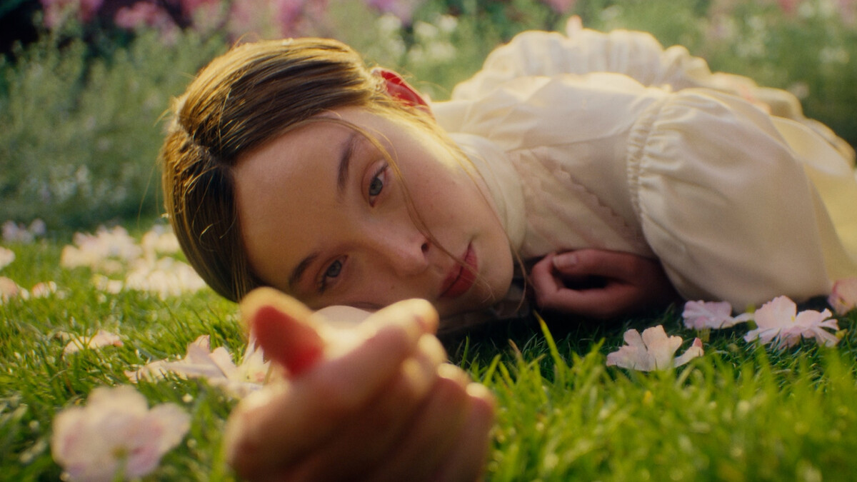 A young woman in a light blouse lies on green grass surrounded by pink flowers, looking directly at the camera in a sunlit garden or field.