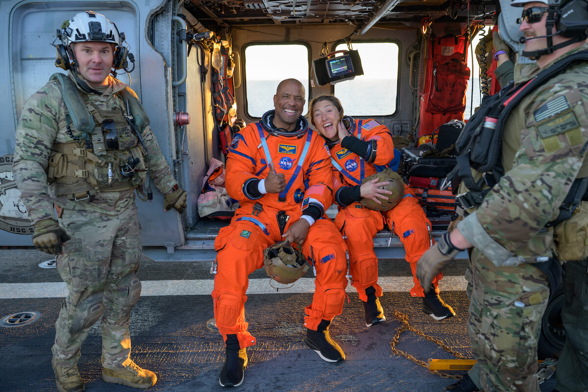Two astronauts in orange NASA suits sit smiling in a helicopter, surrounded by military personnel in camouflage uniforms on a sunlit landing pad, suggesting post-mission recovery at sea.