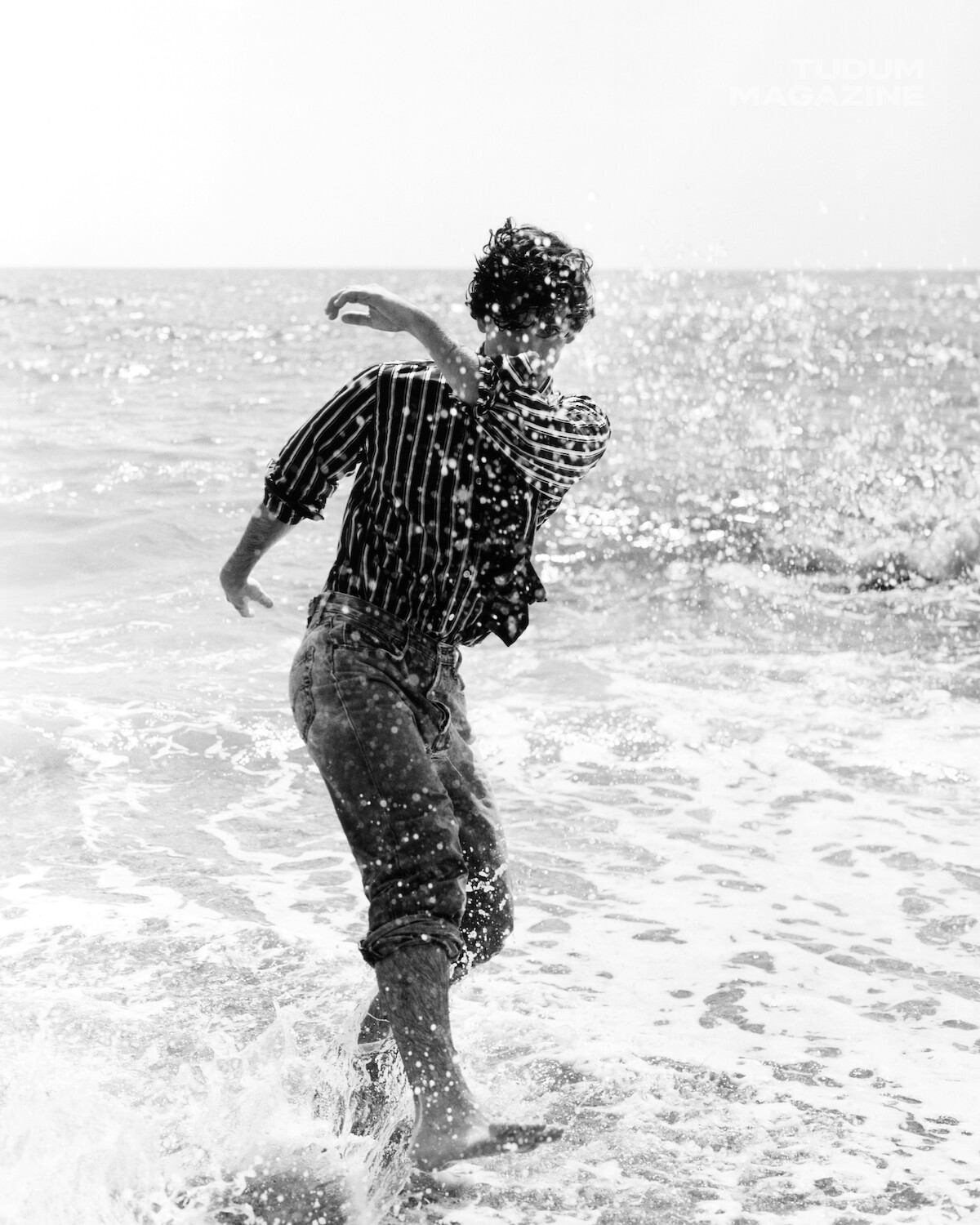 Person in rolled-up jeans and striped shirt playing in ocean waves, splashing water at the beach; shot in black and white with sunlight reflecting on the sea.