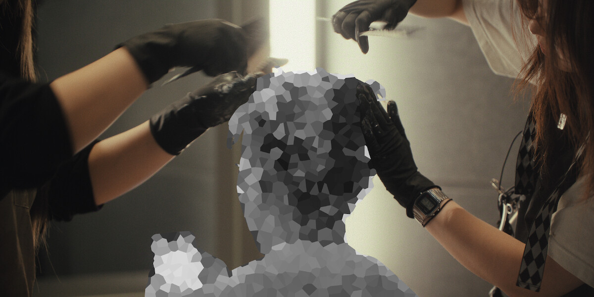 Two people wearing black gloves are styling or cutting a person’s hair in a salon-like setting, but the client’s head and upper body are heavily pixelated, obscuring their features.