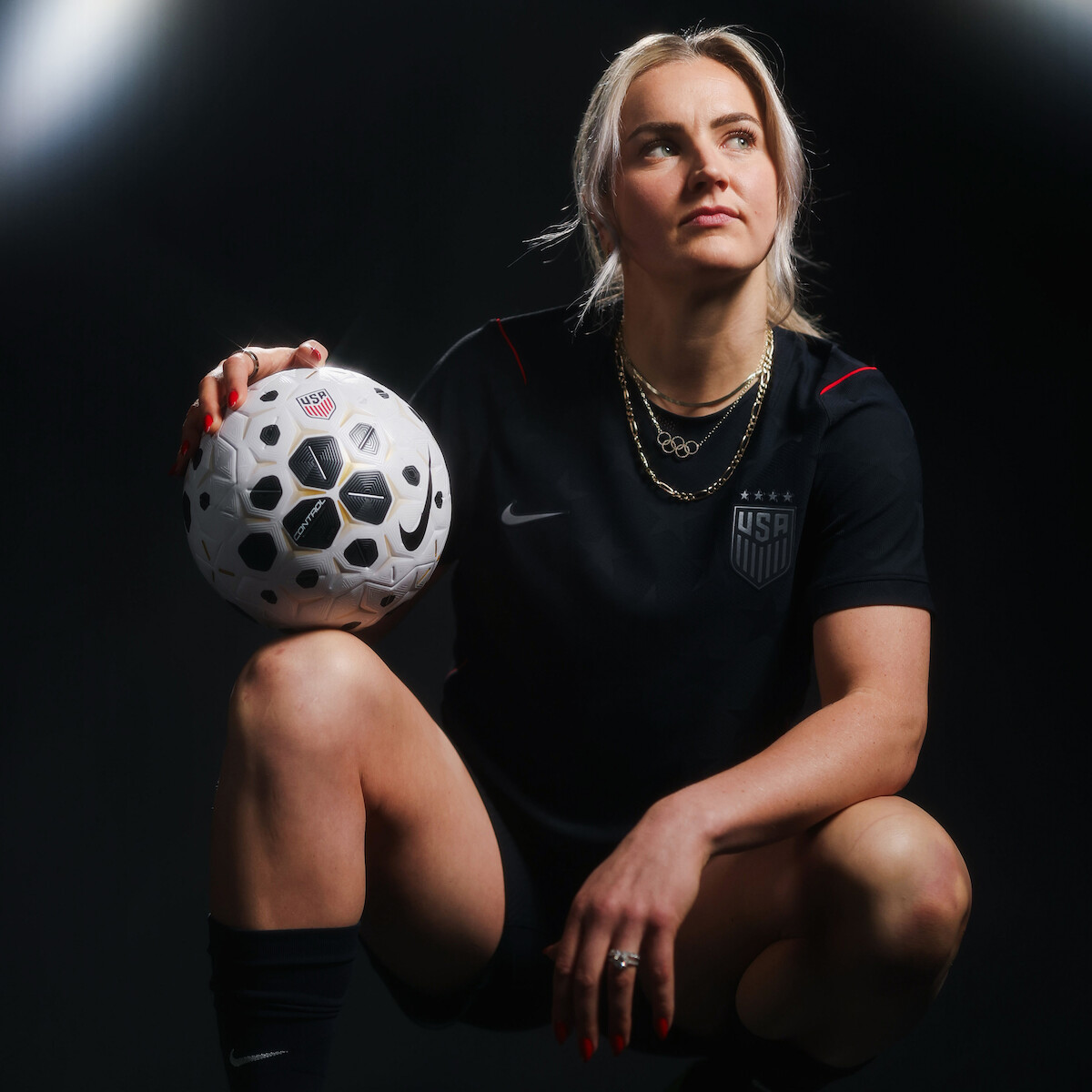 Female soccer player in a black USA uniform poses confidently in a dark studio, kneeling with one hand on her knee and the other resting on a soccer ball, dramatic lighting highlighting her determined expression.