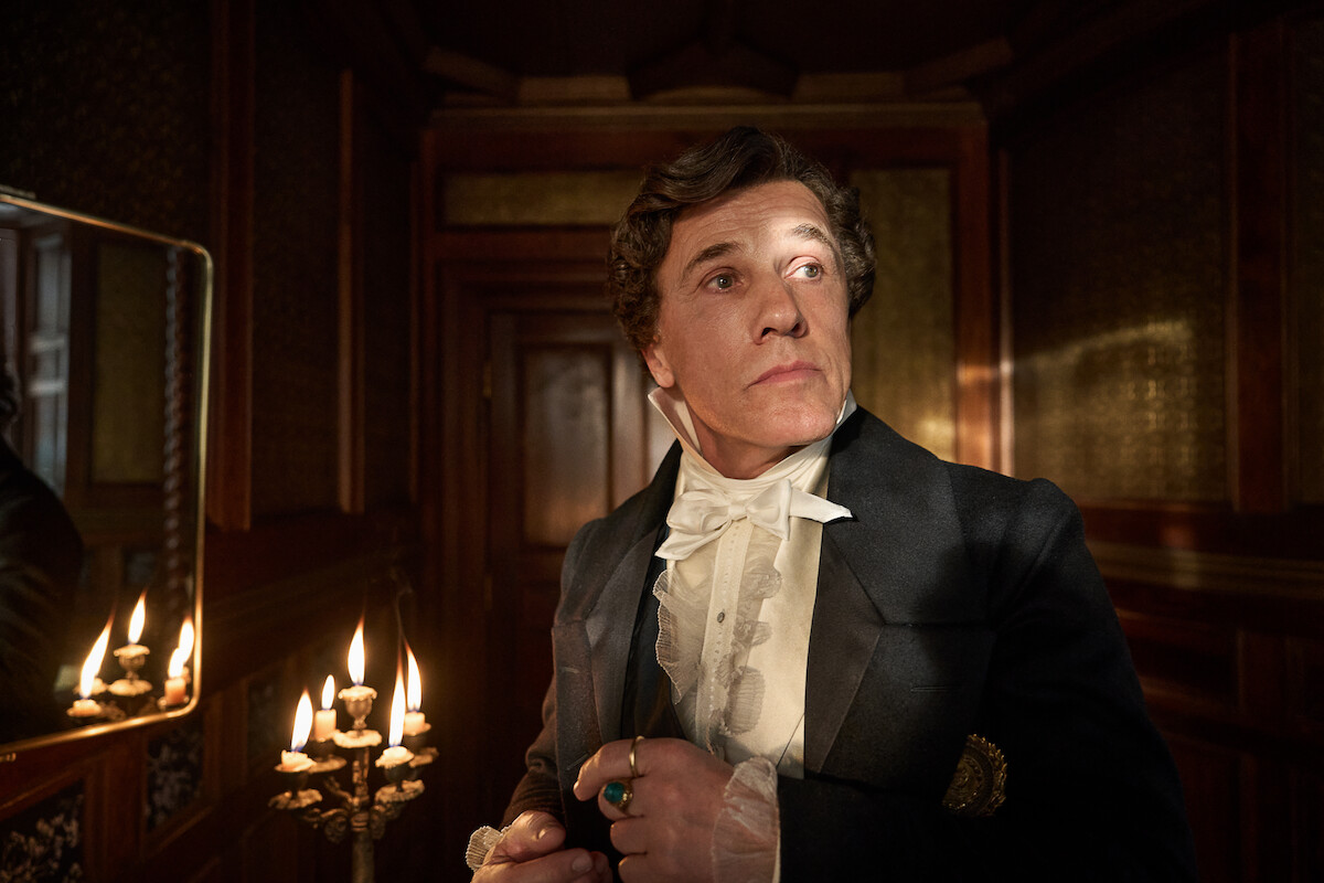 A man in historical attire stands in a dimly lit, ornately decorated room beside a lit candelabrum, looking contemplative and serious, evoking a dramatic, vintage atmosphere.