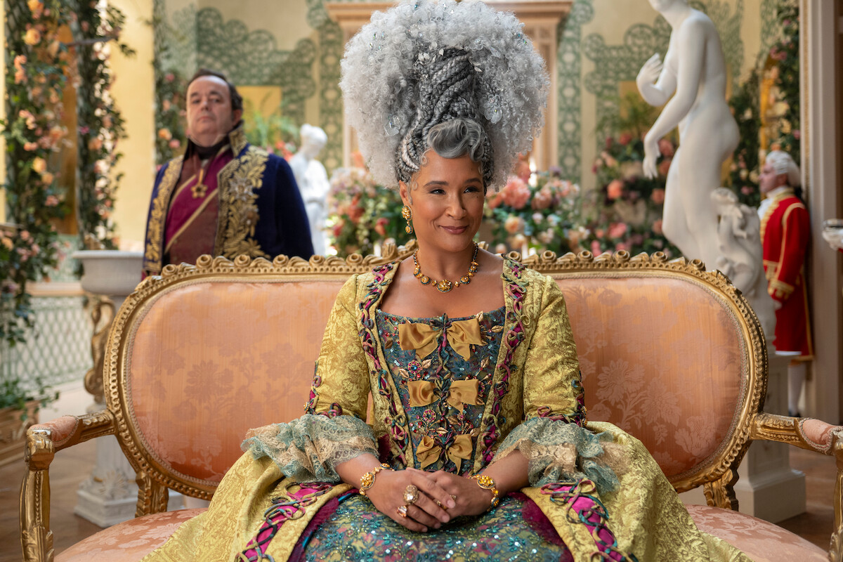 Uma mulher usando um elaborado traje de época está sentada em um luxuoso sofá, em uma sala ricamente decorada com flores e estátuas, com outra pessoa usando trajes históricos ao fundo.