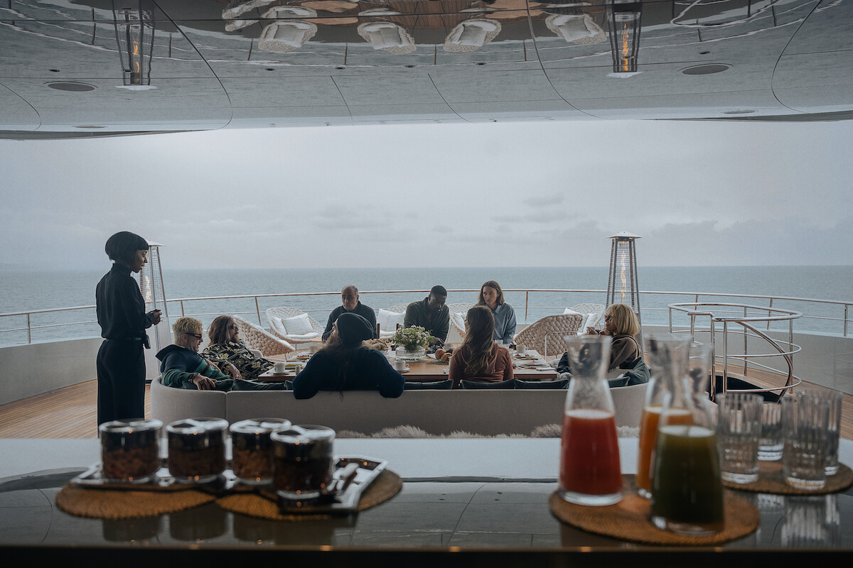 A group of people relax on a yacht deck overlooking the sea on a cloudy day, drinks and juice on a bar in the foreground, creating a calm, luxurious, and social atmosphere.