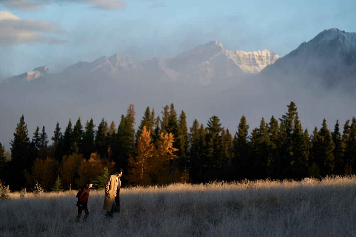 Two people walking through a grassy field at sunrise or sunset with evergreen trees and tall, misty mountains in the background.