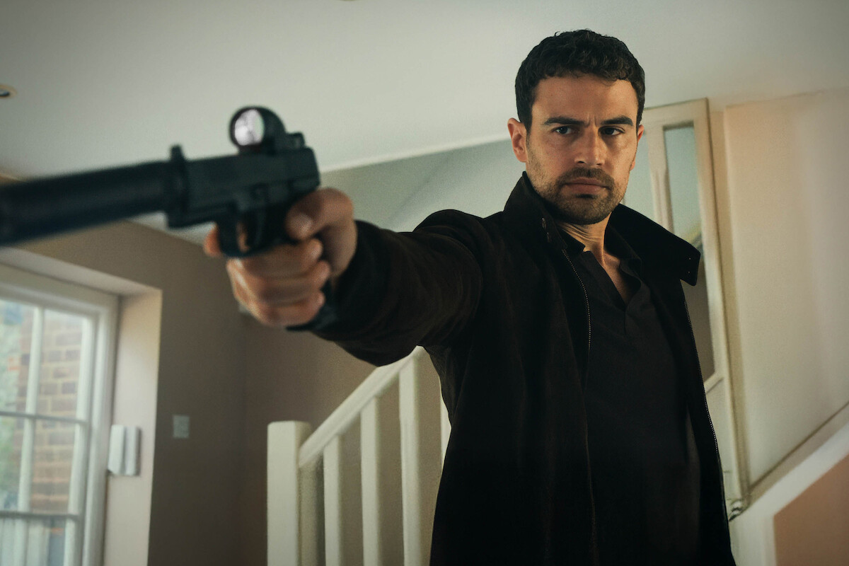 Man indoors in front of a staircase aiming a silenced handgun with a serious expression, wearing a dark jacket, with natural light coming from a window on the left.
