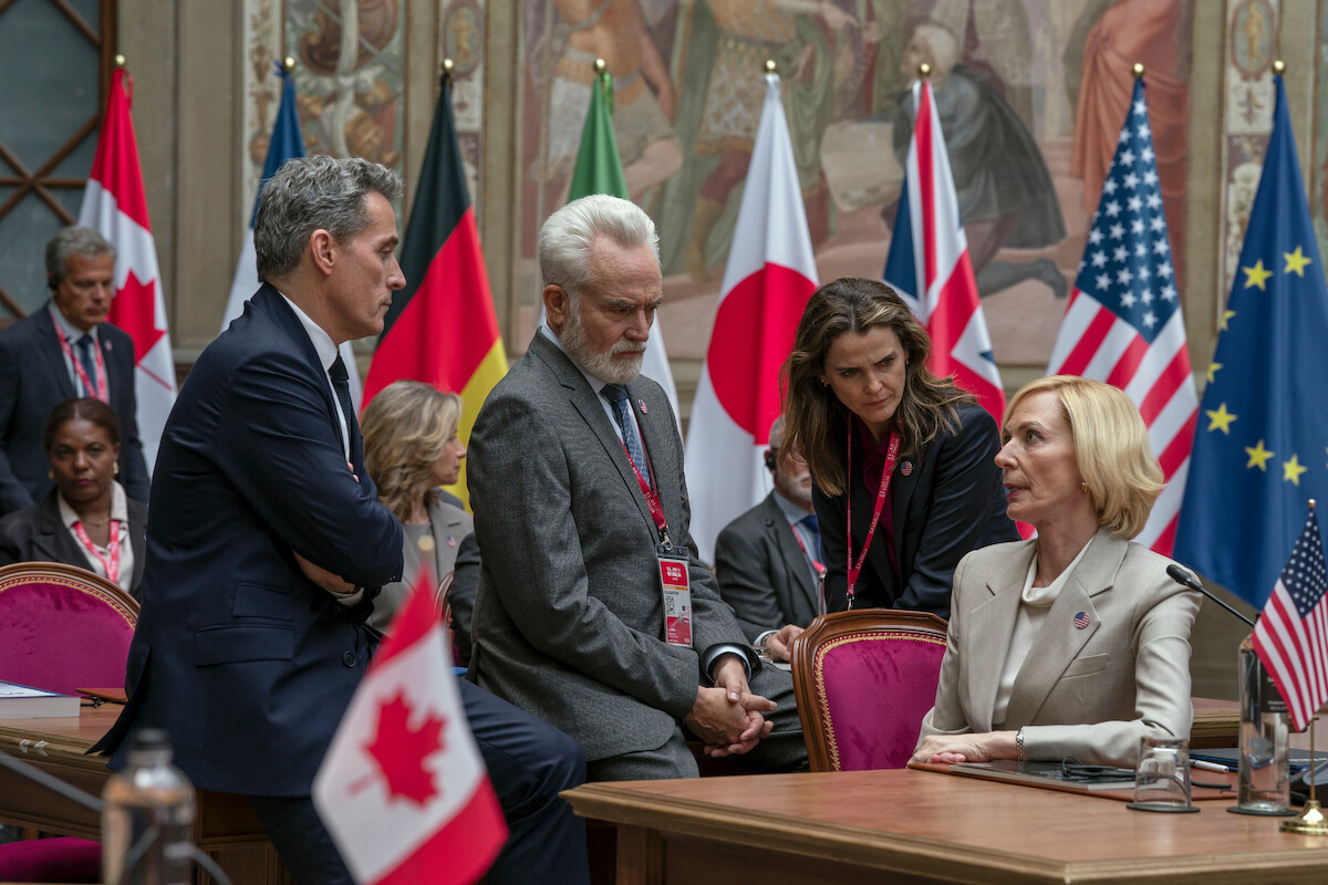 Four people in formal attire have a serious discussion at an international political meeting room, surrounded by flags from various countries including the US, Canada, Japan, and the EU. Conference table with microphones in foreground.