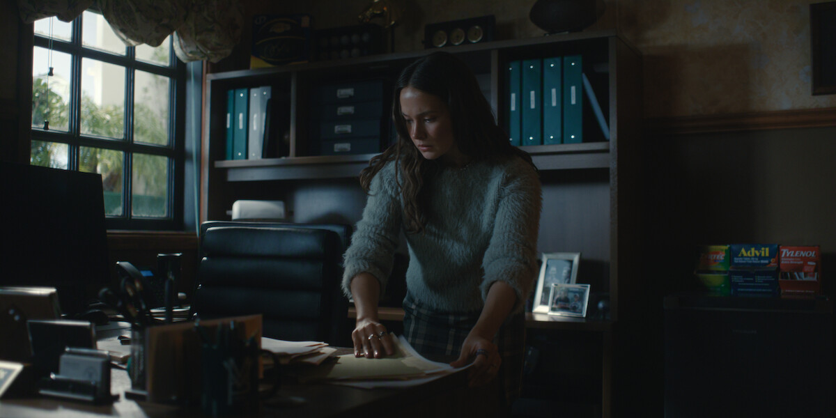 A woman stands at a desk in a dimly lit office, sorting through papers. Bookshelves, framed photos, and medication boxes are visible in the background; daylight is coming in from a window to the left.