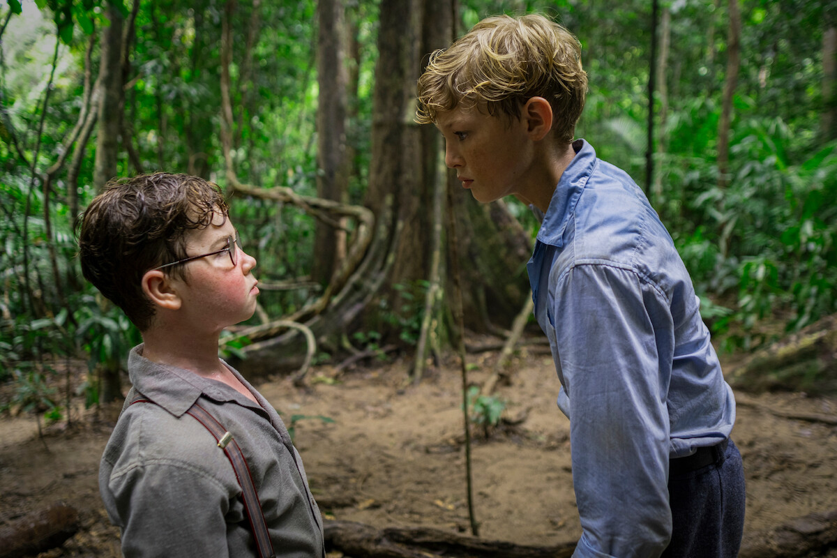 Two boys stand face to face in a tense confrontation in a dense jungle with trees and vines visible in the background, wearing worn clothing and serious expressions, suggesting a dramatic or survival setting.