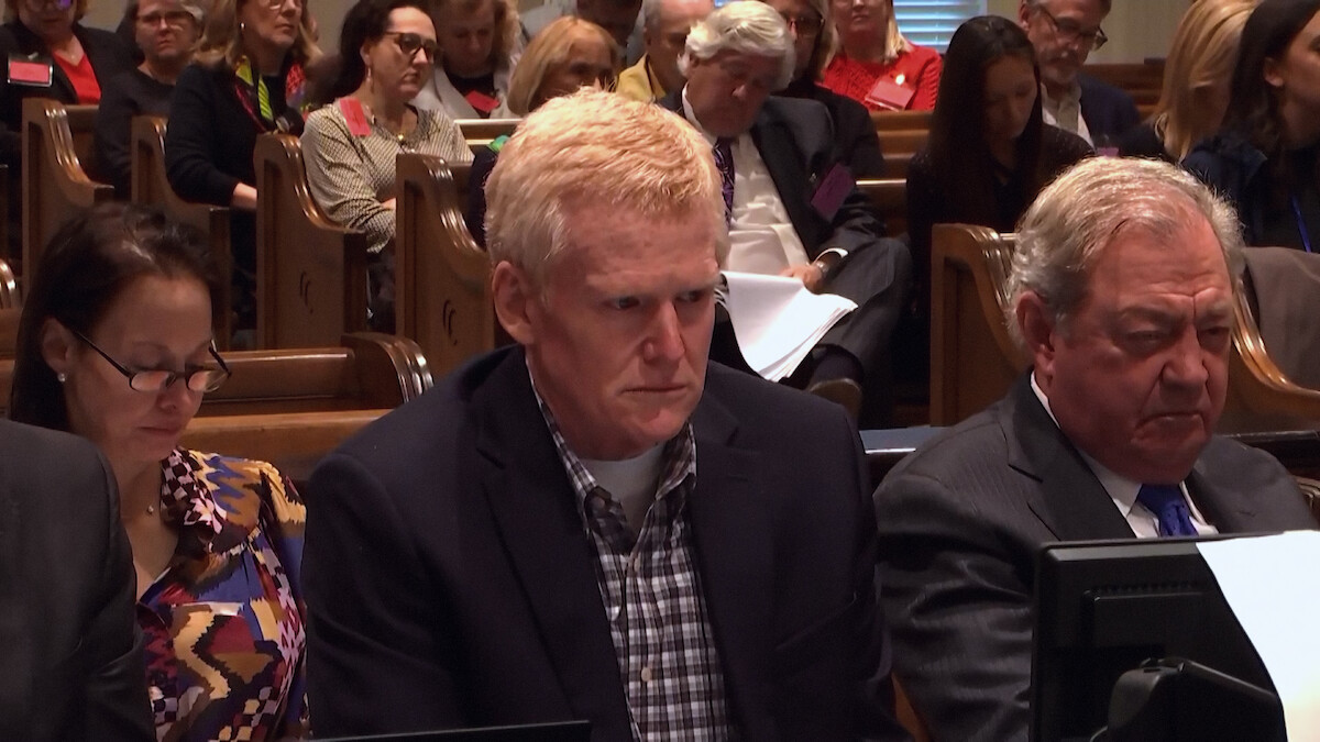 Three people sit at a courtroom table, looking serious. They are surrounded by other people seated in wooden pews, suggesting a legal proceeding in progress. The atmosphere is formal and attentive.