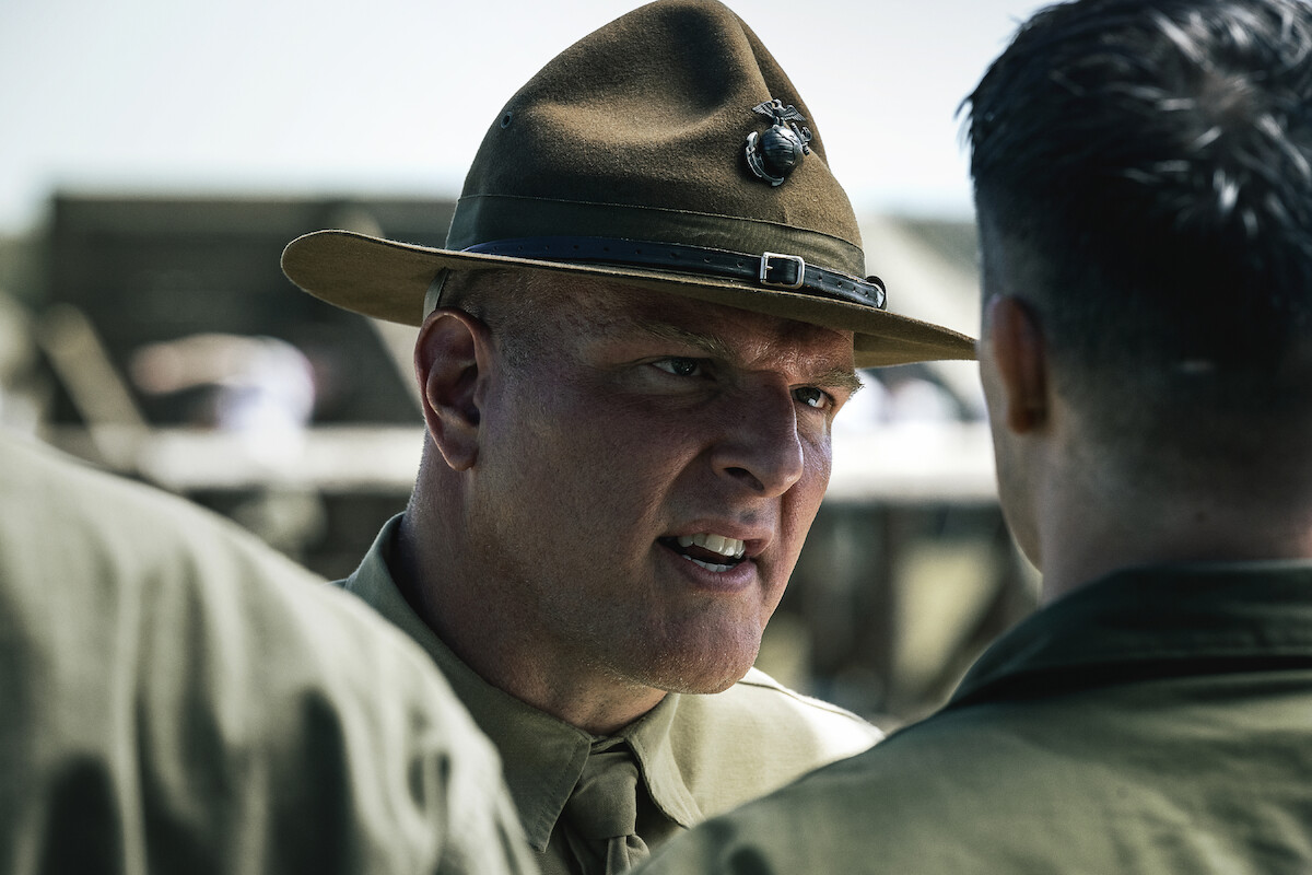 Pat McAfee as Drill Instructor in uniform and campaign hat confronts a recruit outdoors in a training camp, with blurred soldiers and structures in the background under daylight.