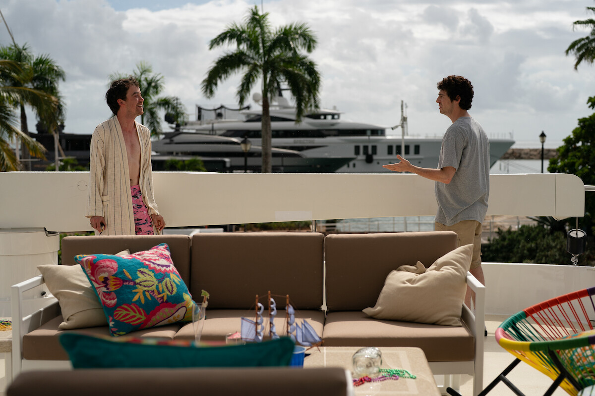 Two people are talking on a sunny outdoor patio with a sofa, colorful pillows, and a table; palm trees and a large yacht are visible in the background, creating a relaxed, tropical setting.