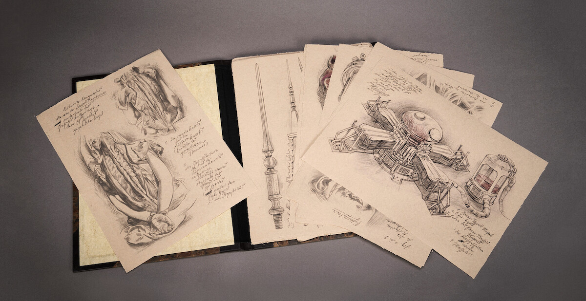 Pencil sketches of intricate fantasy objects and machinery on aged paper, scattered from an open book on a gray surface, evoking a mysterious, creative, and imaginative mood.
