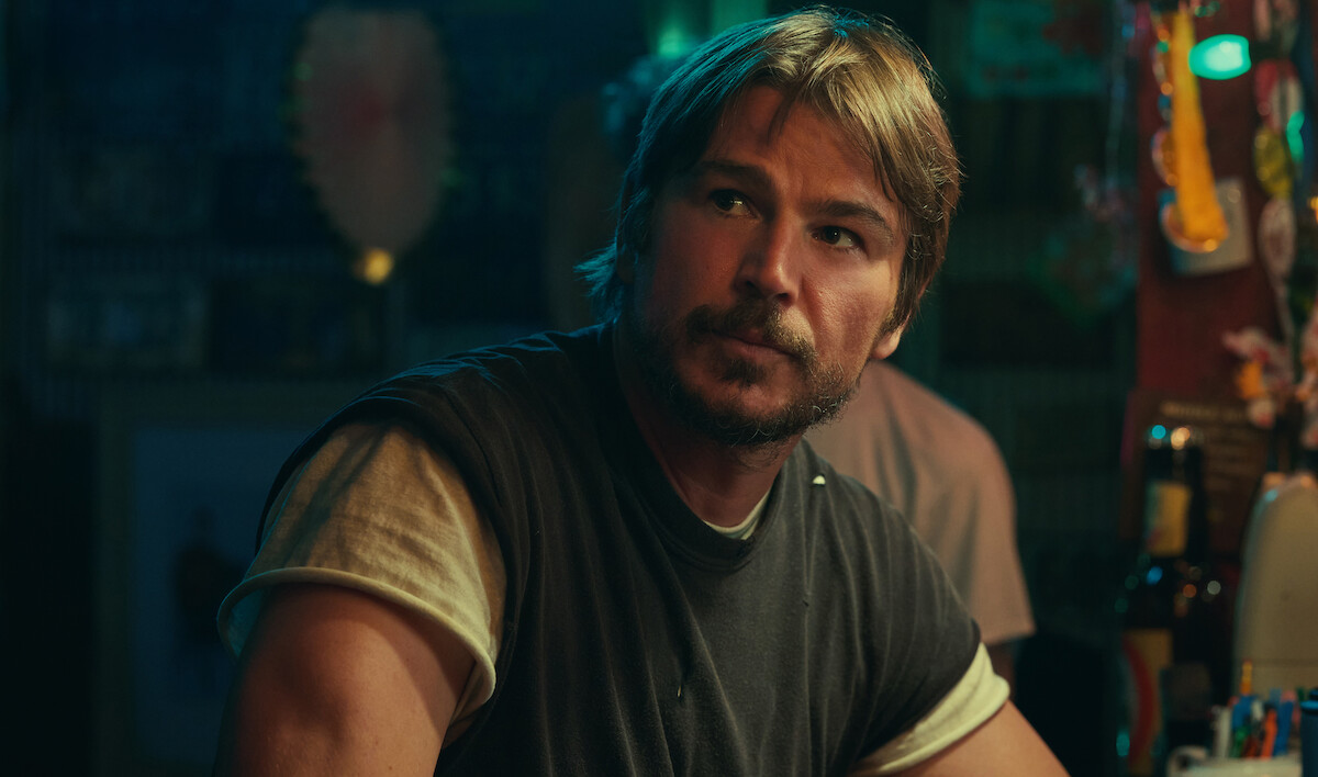 Man with light brown hair and beard sitting at a dimly lit bar counter, wearing a dark t-shirt over a white undershirt, with colorful decorations and bottles visible in the background.
