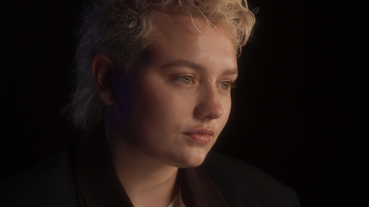 Person with short, light blonde hair in a dark jacket, looking pensive, is softly lit against a dark background in a close-up portrait, creating a calm and introspective atmosphere.