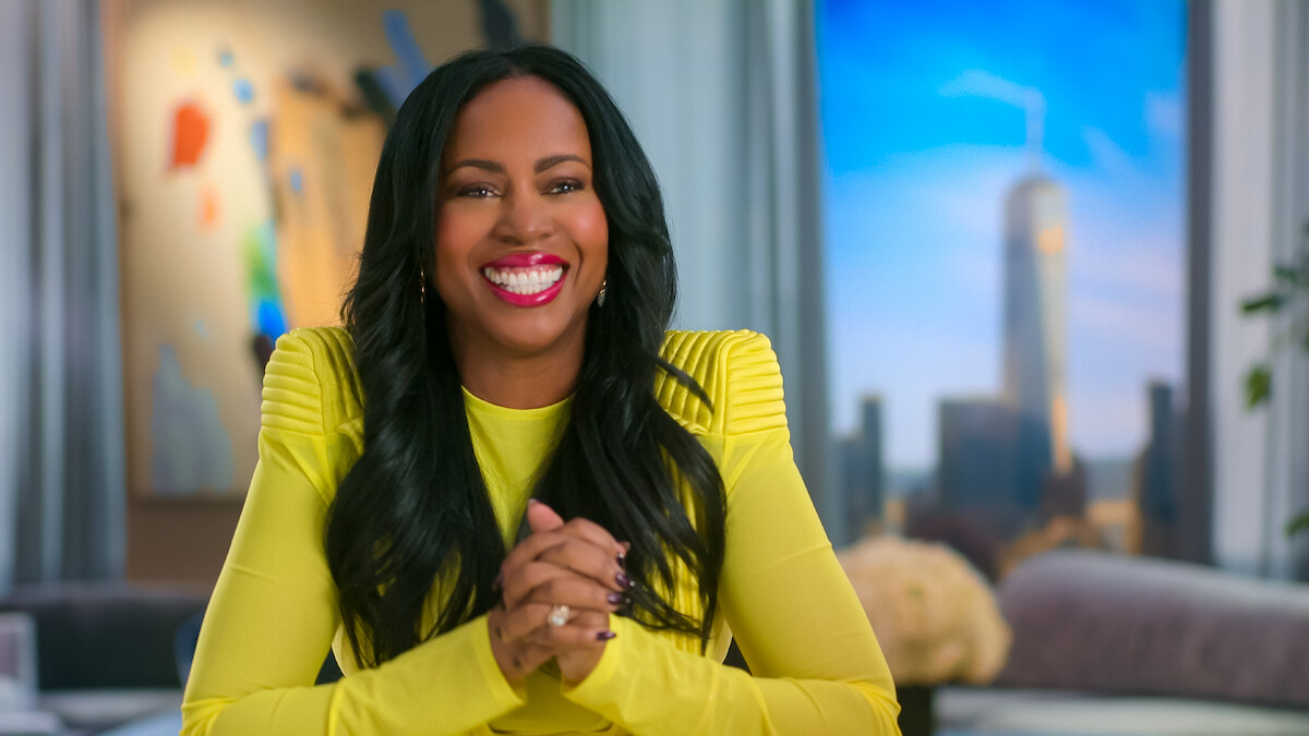 Smiling woman with long black hair in a bright yellow top sits in a modern, sunlit room with abstract art and a cityscape view, creating a cheerful and welcoming atmosphere.