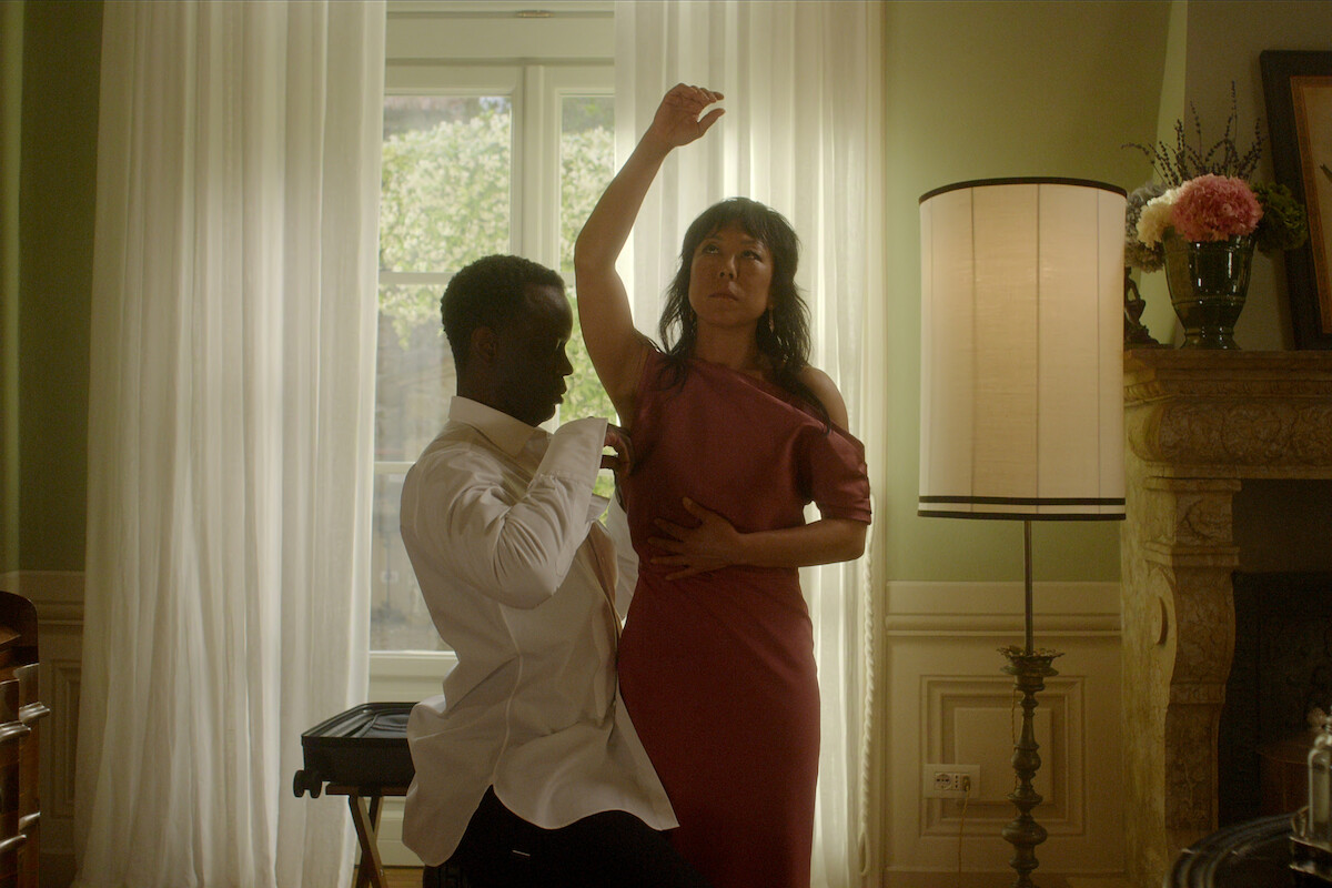 A man in a white shirt kneels and smells a woman's raised arm in a living room with soft natural light, large window, lamp, fireplace, and flowers, creating an intimate and artistic atmosphere.