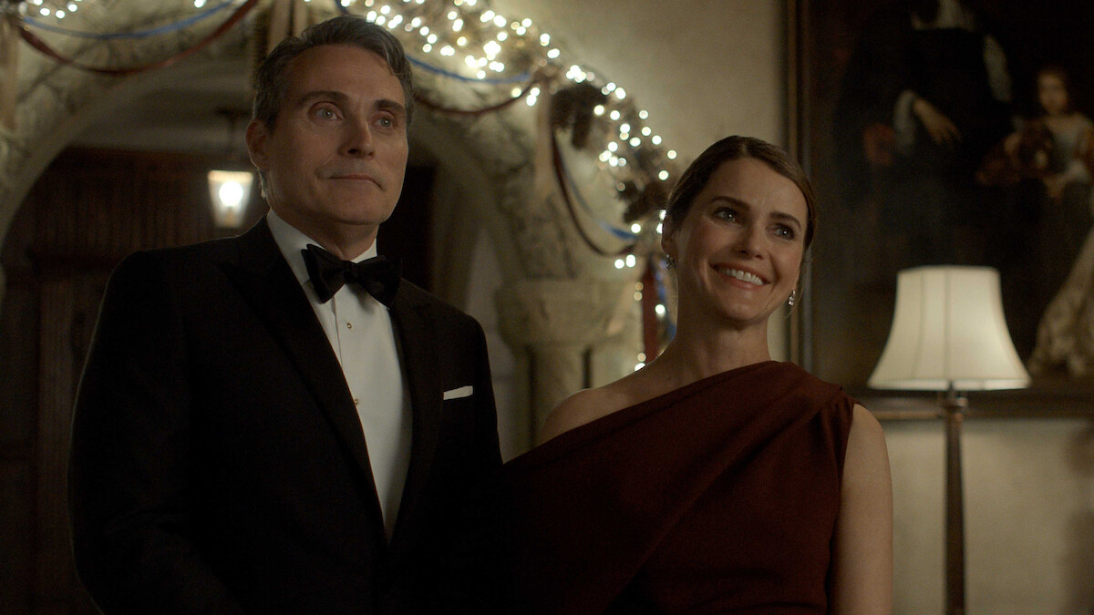 A man in a tuxedo and a woman in an elegant, one-shoulder dress stand smiling together in a warmly lit room decorated with string lights and festive garlands.