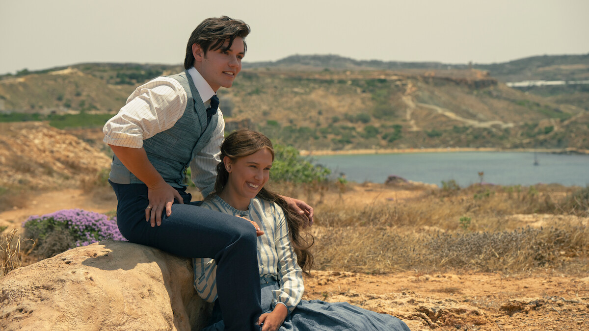 Two people in vintage clothing sit on a rock in a dry, grassy landscape with hills and a body of water in the background, smiling and enjoying the scenic outdoor setting.