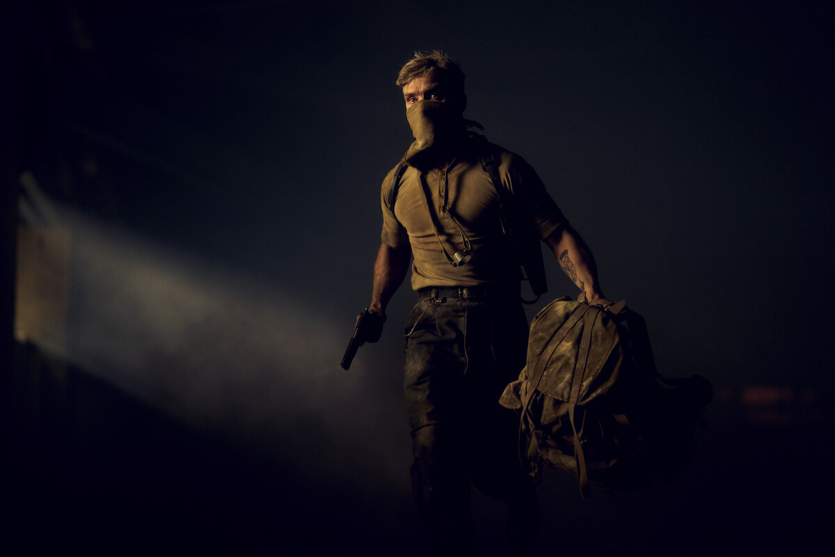 A masked man with a gun and a duffel bag walks through a dark, smoky environment with a beam of light shining in the background, suggesting a tense or action-packed setting.