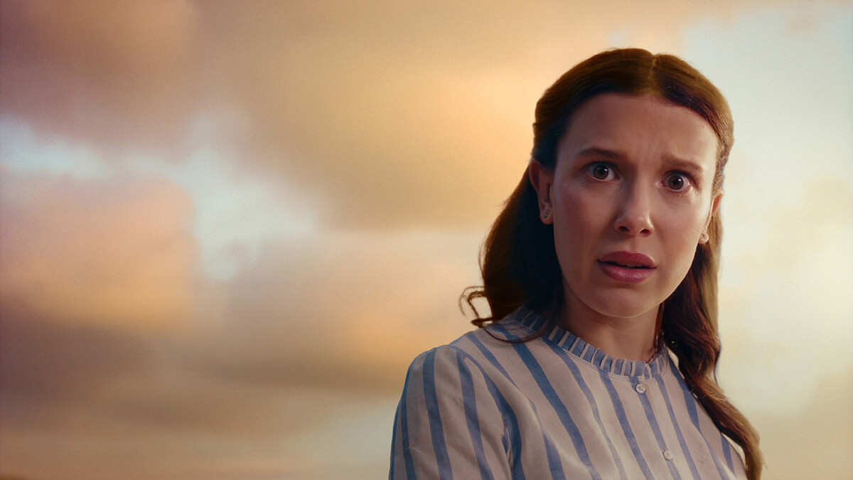 Millie Bobby Brown, wearing a light blue and white striped shirt, looks surprised or concerned outdoors at sunset with a cloudy sky in the background.