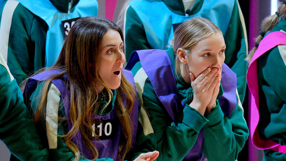 Two women in matching green tracksuits with numbered vests appear shocked and tense, sitting among other similarly dressed people in a brightly lit setting, suggesting a competitive or high-stakes environment.