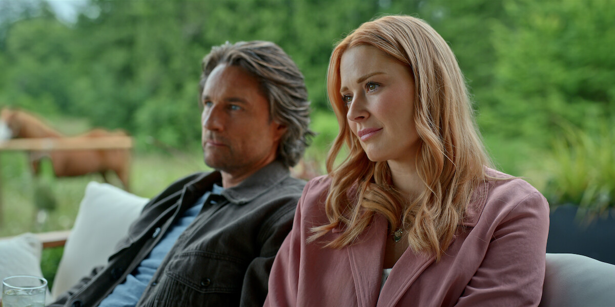 A man and woman sit together on an outdoor sofa with trees and a horse in the background, appearing relaxed in a lush green natural setting.