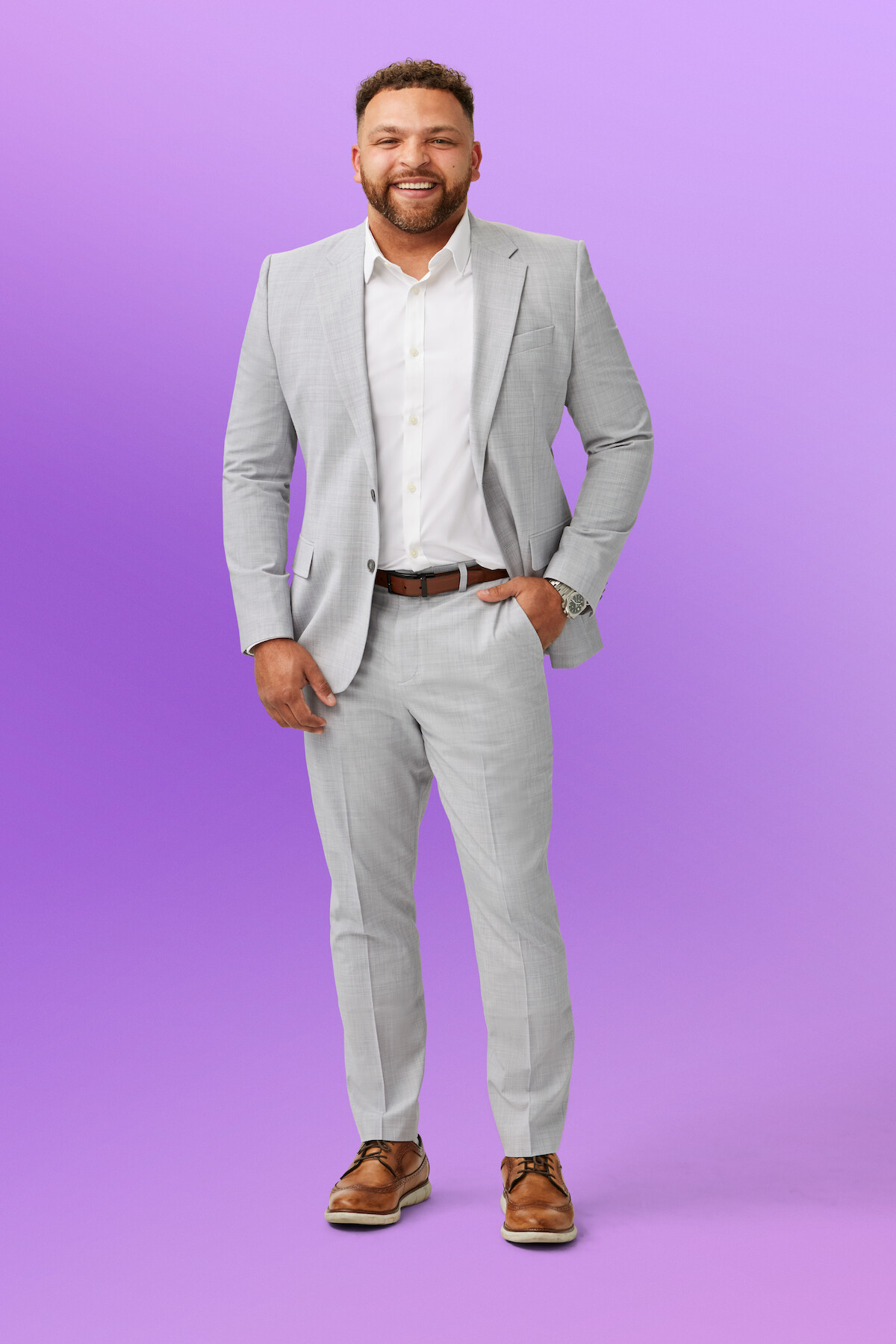 Devonta wearing a light gray suit, white shirt, and brown shoes stands confidently with one hand in his pocket against a solid light purple background in a studio setting.