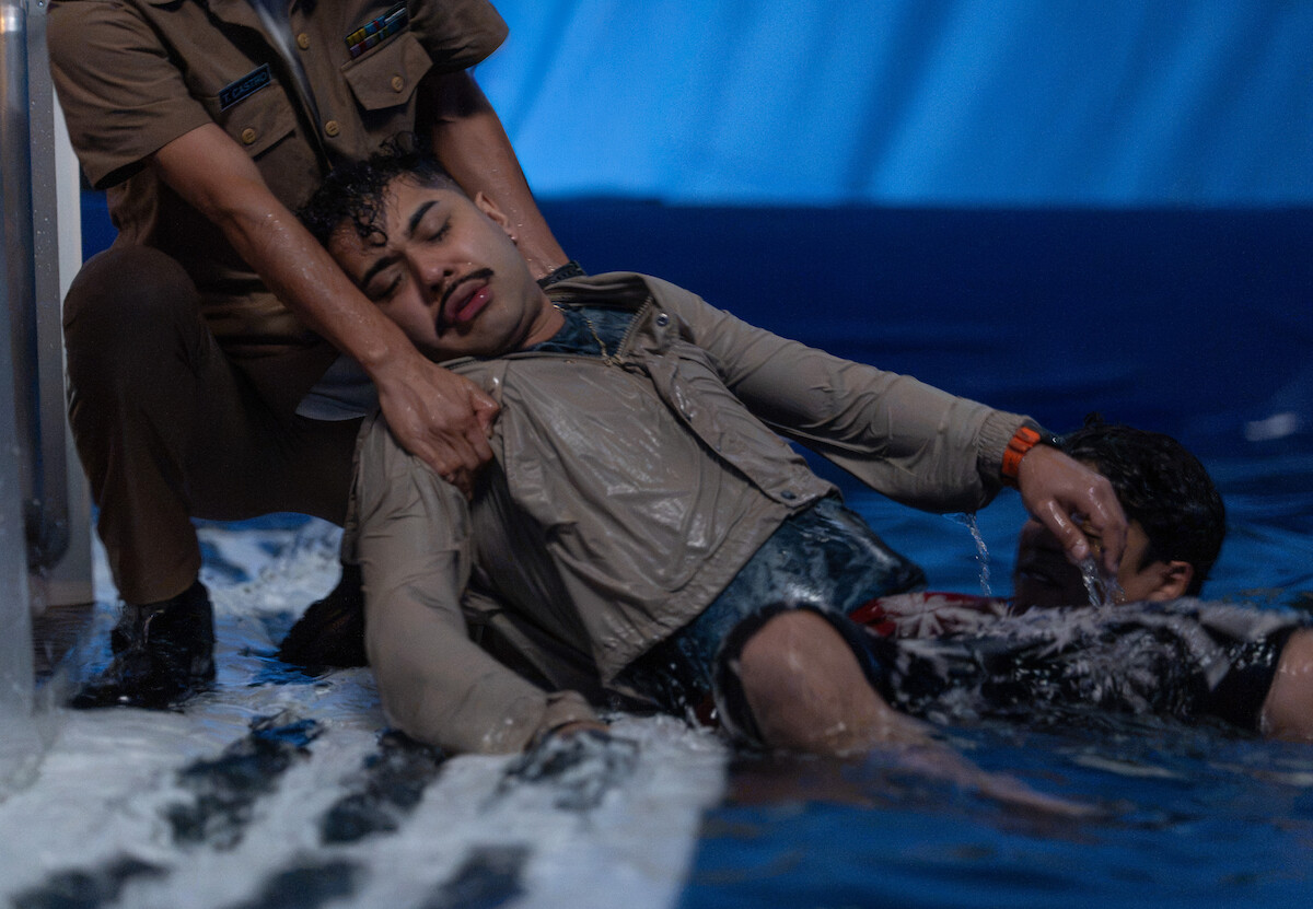 Um homem jovem, de cabelos e bigodes pretos, está sendo retirado da água por outro homem. Ele tem o tronco fora da água, apoiado na parte traseira de um barco, e as pernas ainda submersas. O homem parece estar inconsciente
