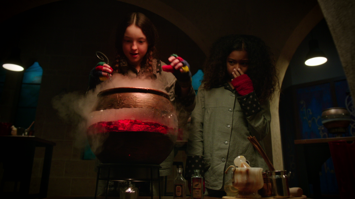 Two girls in a dimly lit room stand over a steaming cauldron, one stirring while the other covers her nose, suggesting a magical experiment or potion-making scene. Various jars and ingredients sit on the table nearby.